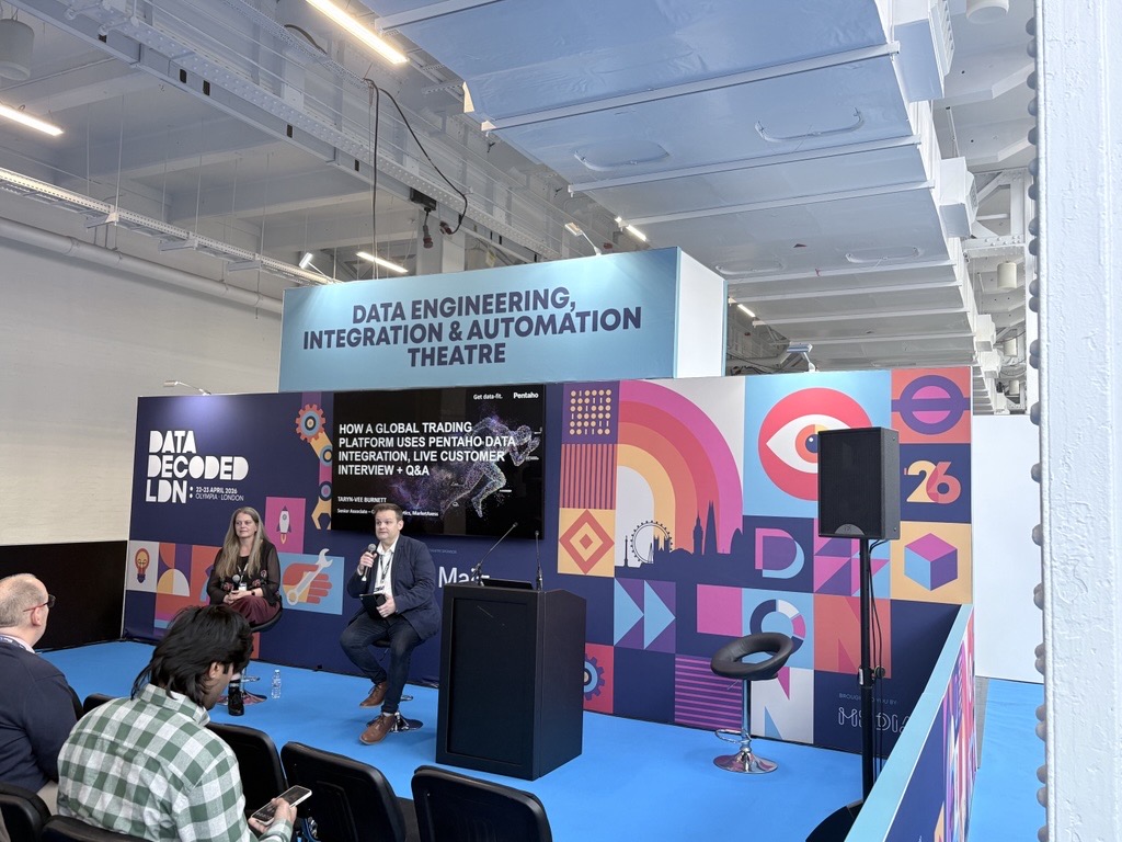 Two speakers sit on a blue stage at a conference, presenting under a banner that reads 'Data Engineering, Integration & Automation Theatre' with a colorful geometric backdrop.